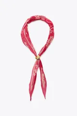 Pañuelo o bandana de seda y algodón, color fucsia intenso, con estampado floral y paisley en tonos beige/crema. Se anuda al cuello con un broche metálico en forma de corazón dorado y verde.