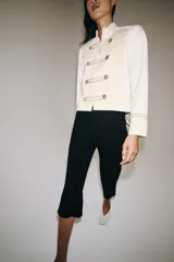 Chaqueta de estilo militar de ZARA Woman Collection, confeccionada en mezcla de algodón en color blanco y beige. Presenta cuello subido, manga larga con puños anchos en contraste y cierre frontal con alamares y botones metálicos en relieve. Tiene bajo asimétrico con abertura en la espalda.