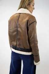 Campera estilo aviador de cuero sintético marrón con interior y detalles de corderito sintético en color crudo. Cuenta con cierre frontal metálico, detalles de tiras con hebilla en el cuello y el ruedo, y ribetes en color negro.