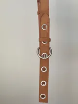 Cinturón de símil gamuza color tostado, con diseño de ojales metálicos plateados a lo largo de toda la pieza y hebilla redonda metálica.
