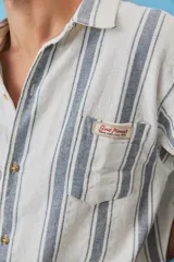 Camisa de manga corta a rayas verticales en tonos azul y blanco, con cuello camisero, cierre frontal de botones y un bolsillo en el pecho con etiqueta aplicada.