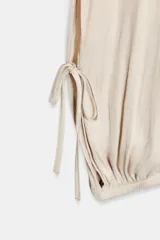 Pantalón de mujer color beige claro, de tiro medio y calce holgado, confeccionado en tejido ligero y fluido. Presenta cintura elástica ajustable con lazada, bolsillos laterales y aberturas ajustables con lazadas en las piernas que terminan en bajo abullonado con elástico.