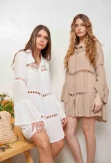 Vestido corto de corte evasé color beige, con cuello camisero, botonadura frontal completa y mangas largas acampanadas. Presenta detalles de bandas con calados circulares en el pecho, mangas y a lo largo de la falda en niveles.