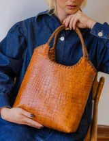 Bolso tipo matera de cuero con textura de reptil, color tostado, con manijas trenzadas y detalles de costura artesanal en los bordes.