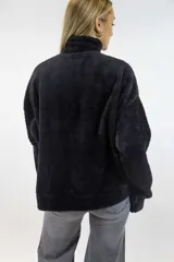 Buzo de peluche color blanco roto, con cuello alto, cierre frontal parcial, mangas largas y un bolsillo tipo canguro al frente.