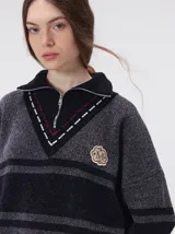 Pullover de punto con textura aterciopelada, diseño oversize, cuello polo con cierre frontal parcial y detalles de pespuntes en contraste. Presenta rayas horizontales en tonos tierra y un parche bordado con la letra M en el pecho.