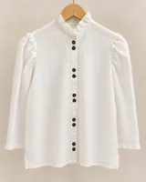 Camisa blanca de lino, con cuello alto con volados, mangas largas abullonadas en los hombros y cierre frontal con botones oscuros.