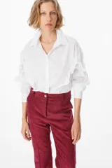 Camisa de corte cropped en color azul, con cuello clásico y cierre frontal de botones. Presenta volados decorativos que recorren la parte frontal y las mangas largas, terminando en puños abotonados.