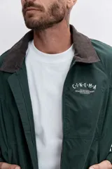 Cazadora de estilo urbano en color verde oscuro, con cuello de pana en tono marrón, cierre frontal con cremallera y puños elásticos. Presenta un diseño holgado con bordado pequeño en el pecho.