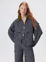 Campera Mutma de denim grueso con diseño a rayas en tonos gris y blanco, corte oversize, cuello solapa de pana verde militar y cierre frontal de cremallera doble. Cuenta con mangas desmontables mediante cierre en los hombros, canesú en el pecho y bolsillos de parche delanteros.