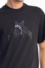 Remera de algodón color negro, con cuello redondo y mangas cortas. Presenta un estampado frontal con una estrella azul y un delfín en el centro, acompañado de tipografía estilo gótico.