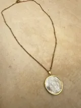 Collar de cadena dorada con eslabones pequeños y detalles de esferas, que sostiene un dije circular con la imagen de la Virgen tallada sobre una base de nácar y borde metálico dorado.