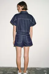 Camisa corta de denim de manga corta, con cuello solapa, escote en V y bolsillos delanteros de plastrón. Cuenta con costuras en contraste y cierre frontal con botones metálicos.