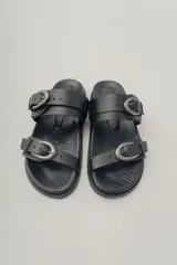Sandalias planas de cuero negro con diseño de dos tiras ajustables con hebillas metálicas.