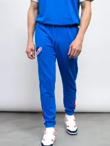 Pantalón de jogging azul de felpa, con detalles de piping y franjas laterales en rojo y blanco. Incluye el escudo del Club Nacional de Football en el muslo derecho.
