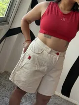 Bermuda de jean color crudo con costuras y detalles en rojo, incluyendo un botón frontal y un logo bordado en el bolsillo lateral. Presenta un corte recto y largo hasta la rodilla.