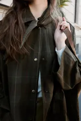 Chaqueta de algodón encerado con diseño de cuadros en tonos verde oliva, beige y líneas rojas. Presenta cuello solapa, cierre frontal con botones a presión metálicos, bolsillos de plastrón con solapa y un detalle de pañuelo desmontable sobre los hombros.