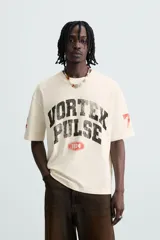 Camiseta de manga corta color crema, corte boxy fit, con cuello redondo. Presenta un estampado frontal en negro con el texto 'VORTEX PULSE' y el número '1974' en un óvalo rojo desgastado. Tiene pequeños detalles gráficos rojos en las mangas.