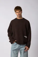 Sweater de punto color marrón oscuro, con diseño de cuello redondo, corte oversize y detalle de bordado pequeño en la parte inferior frontal.