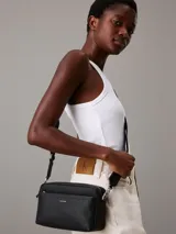 Cartera tipo cámara convertible Calvin Klein negra, con textura monograma en el bolsillo frontal y logo metálico plateado. Incluye dos correas desmontables: una ancha ajustable para cruzar y una fina para llevar al hombro.