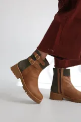 Bota estilo biker de caña media, confeccionada en cuero negro con detalles en gamuza marrón. Presenta correas ajustables con hebillas metálicas, tachas decorativas y suela track dentada.