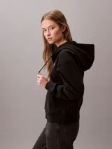Buzo con capucha y cierre frontal de cremallera de la marca Calvin Klein, color negro, de corte regular cropped y manga larga. Presenta bolsillos tipo canguro, cordones ajustables en la capucha y el logo monograma de la marca estampado en el pecho.