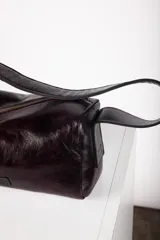 Cartera tipo baguette confeccionada en cuero de color marrón oscuro, con diseño alargado, cierre diagonal y correa de hombro.
