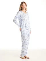 Conjunto de pijama de dos piezas compuesto por una remera de manga larga con escote en V, botones frontales y bolsillo aplicado, y un pantalón largo con cintura elástica y lazo de ajuste. Presenta un estampado floral en tonos celestes y rosados sobre fondo blanco.