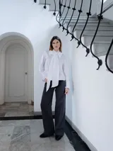Camisa blanca de corte clásico con cuello alto y detalle de lazo grande anudado al frente. Posee mangas largas con puños abotonados y un diseño de calce recto.
