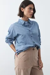 Camisa de jean en tencel con silueta amplia, hombros caídos, cuello clásico y cierre frontal con botones. Presenta ruedo curvo con tajos laterales y pliegue en la espalda.