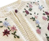 Cardigan de punto color marfil, con cuello redondo a la base, mangas largas y cierre frontal con botones. Presenta un delicado bordado artesanal de flores en tonos bordó, rosado y celeste con tallos y hojas verdes en el frente y las mangas.