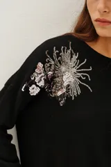 Sweater de punto negro con cuello redondo y mangas largas. Presenta un detalle bordado con lentejuelas en forma de hojas y flores en el hombro derecho.