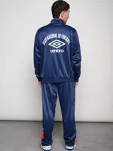 Conjunto deportivo Umbro del Club Nacional de Football, compuesto por una campera con cierre frontal y pantalón a juego. La campera es de color azul con detalles en rojo en los hombros y líneas blancas, presentando el logo de la marca y el escudo del club en el pecho. El conjunto está confeccionado en tejido sintético con acabado satinado.