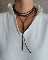 Collar estilo corbatín confeccionado en tiento de cuero marrón, con múltiples cuentas metálicas decorativas distribuidas a lo largo del diseño.