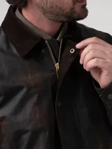 Chaqueta encerada de estilo clásico, color marrón oscuro, con cuello de cotelé y cierre frontal mediante broches a presión. Presenta un diseño de corte regular con bolsillos laterales integrados.