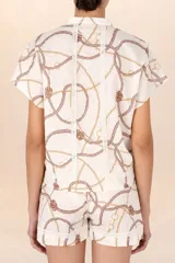 Camisa blanca con estampado de cadenas y cuerdas en tonos dorados y marrones. Tiene cuello mao, mangas cortas y detalles de encaje en el frente y la espalda.