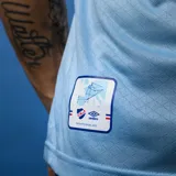 Camiseta de fútbol celeste del Club Nacional de Football, marca Umbro, con cuello en V y detalles en blanco y rojo. Incluye escudo del club, logo de Umbro y sponsors.