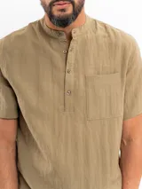 Camisa de manga corta color beige, con cuello mao, bolsillo ojal en el pecho y cierre frontal parcial con botones.