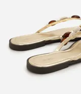 Sandalias planas tipo mule de dedo de la marca Satinato, en color dorado con plantilla metalizada y suela baja. Presenta una tira con aplicaciones decorativas de cuentas de resina en tonos marrones y marfil.