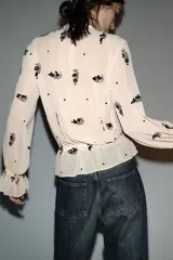 Blusa de cuello subido y manga larga con puños acampanados elásticos. Confeccionada en tejido semitransparente color beige con bordado de lentejuelas oscuras. Presenta cierre en la espalda con abertura y botones ocultos.