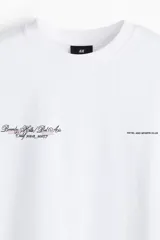 Remera de punto de algodón color blanco, con cuello redondo ribeteado, hombros caídos y manga corta. Presenta un pequeño diseño estampado en el pecho con texto en tipografía cursiva.