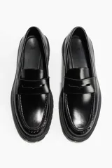 Mocasines de cuero sintético negro con suela gruesa track, costuras prominentes en la puntera y detalle de antifaz en el empeine.