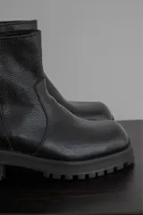 Bota corta de cuero negro con punta cuadrada, suela track y detalle de estribo desmontable con hebilla metálica.