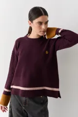 Buzo de punto color crudo con mangas de textura trenzada, detalles en contraste en los puños y franjas horizontales en el ruedo. Presenta cuello redondo y un pequeño bordado en el pecho.