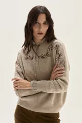 Sweater de tejido de punto color beige con diseño de arabescos en tonos marrones en hombros y mangas. Presenta cuello redondo con cordones decorativos y puños con textura acanalada.