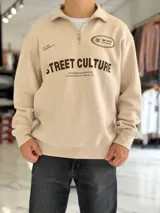Buzo de felpa color beige con medio cierre frontal y cuello alto. Presenta estampado frontal con texto en contraste y diseño de estilo urbano.