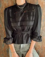 Blusa negra de manga 3/4 con cuello alto, confeccionada en algodón con paneles horizontales de encaje negro en el torso. Presenta un fajín integrado que se anuda a la cintura y puños con volado de encaje.