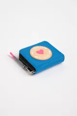 Billetera celeste con cierre de cremallera y tirador de color rosa. Tiene un parche redondo con borde color piel y un corazón rosa en el centro. El parche dice 'Self Love Club' alrededor del corazón.