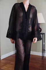 Blusa semitransparente negra de cuello solapa y manga larga, con cierre frontal de botones y detalles de bordados tonales en el ruedo y los puños.