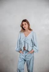 Camisa de tela liviana color celeste, con escote en V y cierre frontal mediante lazos ajustables. Presenta un diseño con volados horizontales en el pecho y mangas largas abullonadas con puños elásticos.